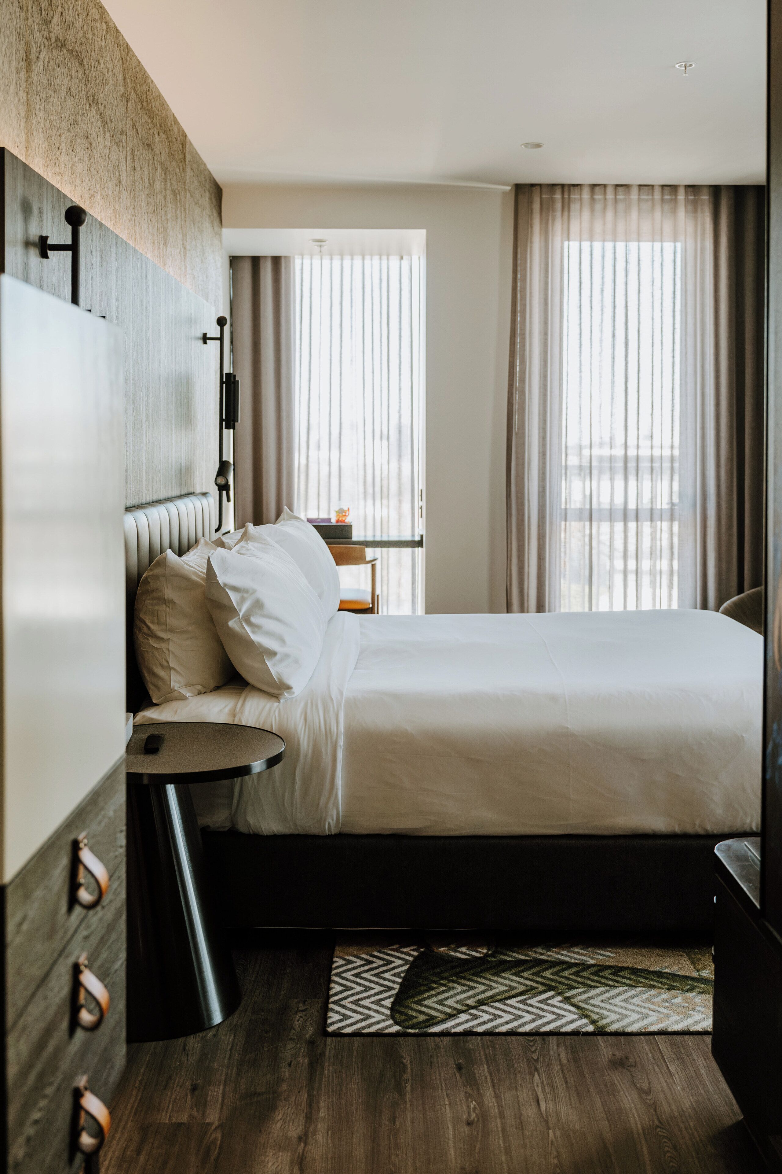 A modern hotel room with a large bed, a bedside table, a lamp, and a view of the city through the window.
