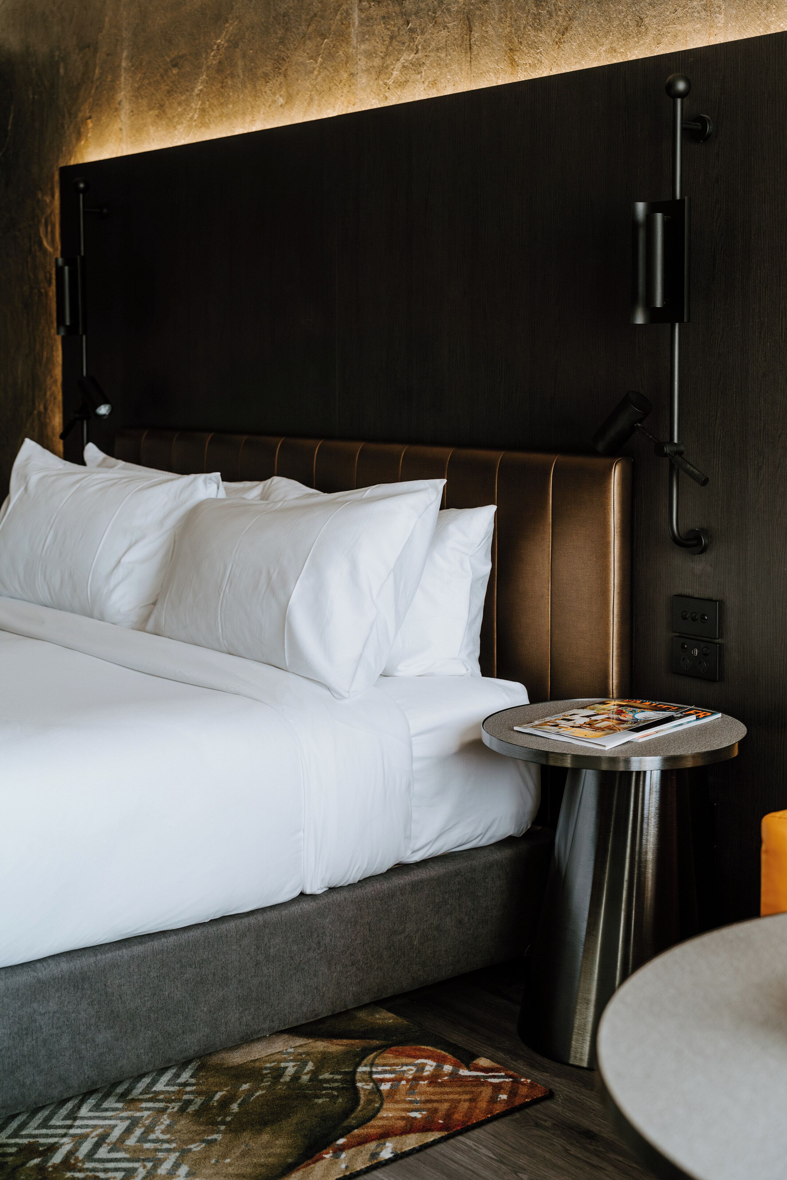 A neatly made bed with white linens, a round side table with a magazine, and a modern headboard.