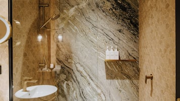 A bathroom with a marble wall, a gold shower, and a white sink.