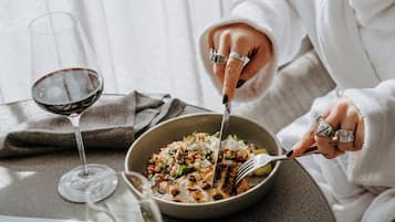 A person in a white bathrobe sitting at a table with a bowl of food, a glass of red wine, and a glass of white wine.