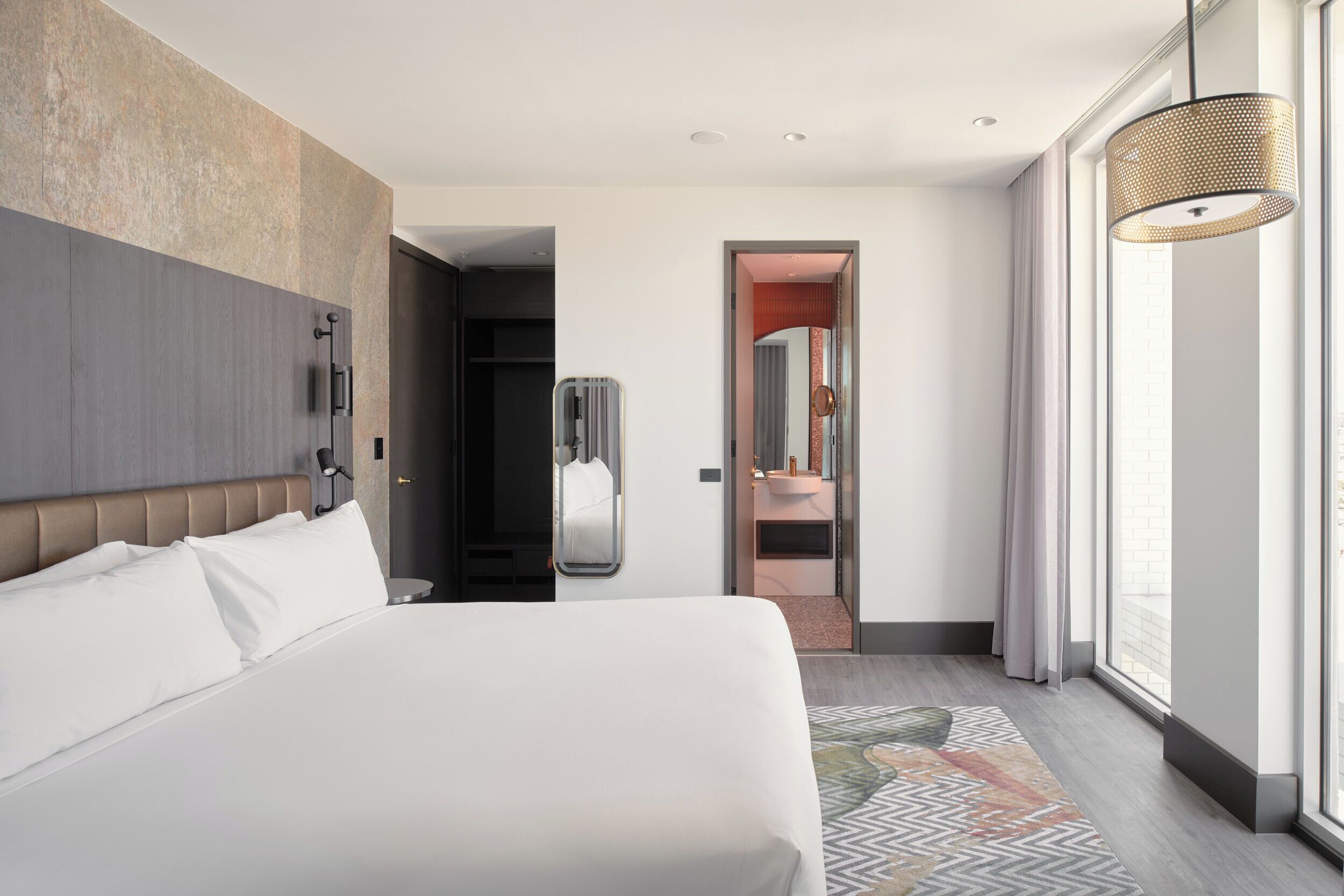 Ett modernt hotellrum med en stor säng, en rymlig klädkammare, ett badrum med spegel och ett stort fönster med gardiner.