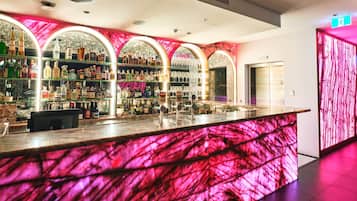 Una zona de bar con mostrador de mármol, estantes llenos de botellas de licor y una pared iluminada en rosa.