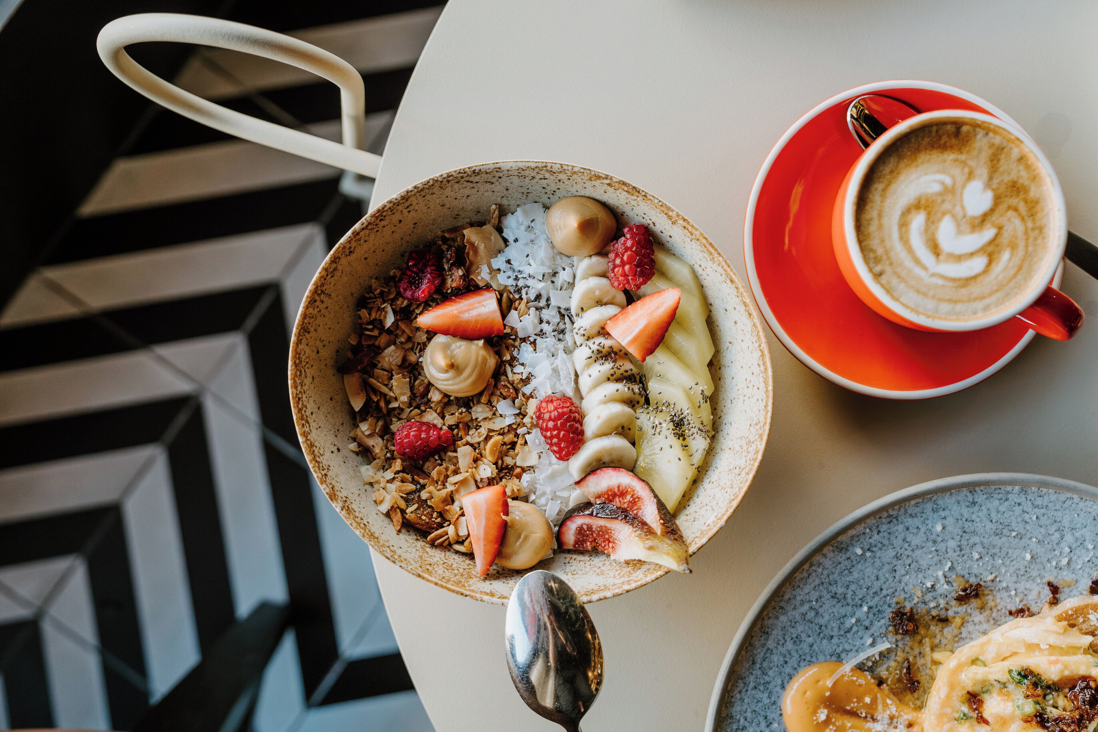A breakfast setting with a bowl of cereal topped with fruits and granola, a cup of coffee with latte art, and a plate with a dessert.
