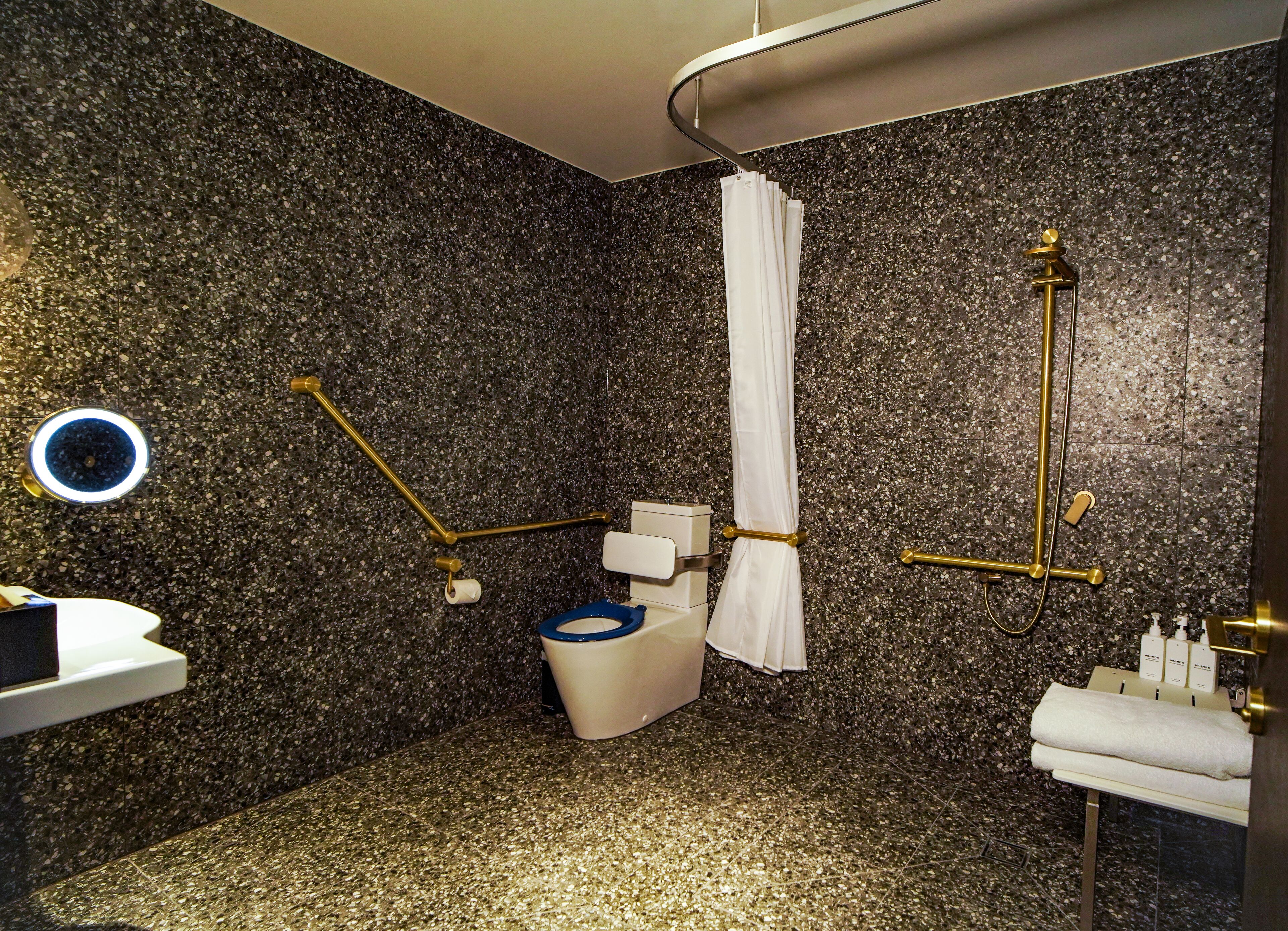 Une salle de bain moderne avec un mur en granit, des toilettes avec un abattant bleu, une douche avec un rideau blanc et un lavabo avec un robinet doré.