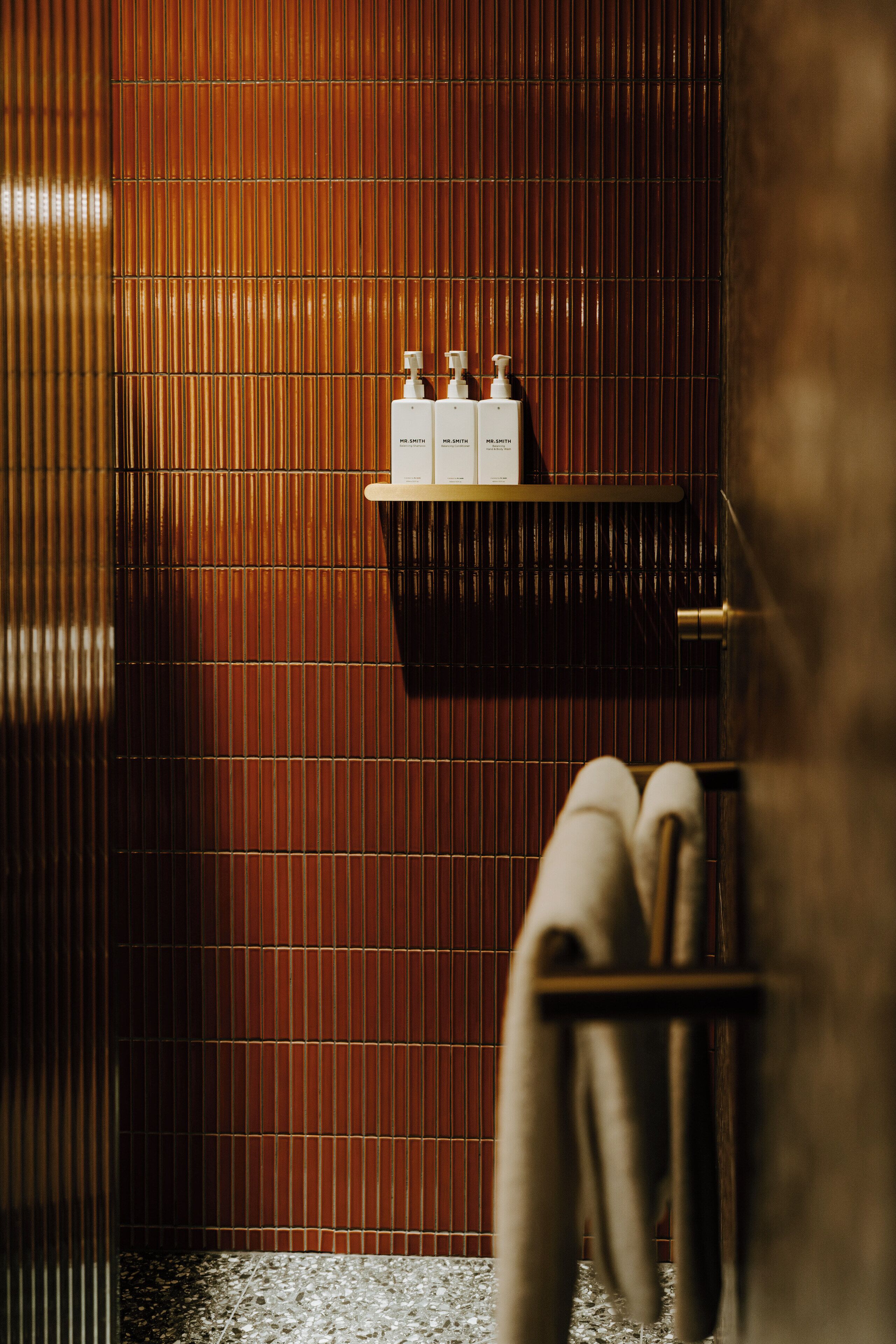 Une salle de bain avec un mur à la texture de bambou, un porte-serviettes et une étagère sur laquelle sont posées trois bouteilles étiquetées « M. Smith ».