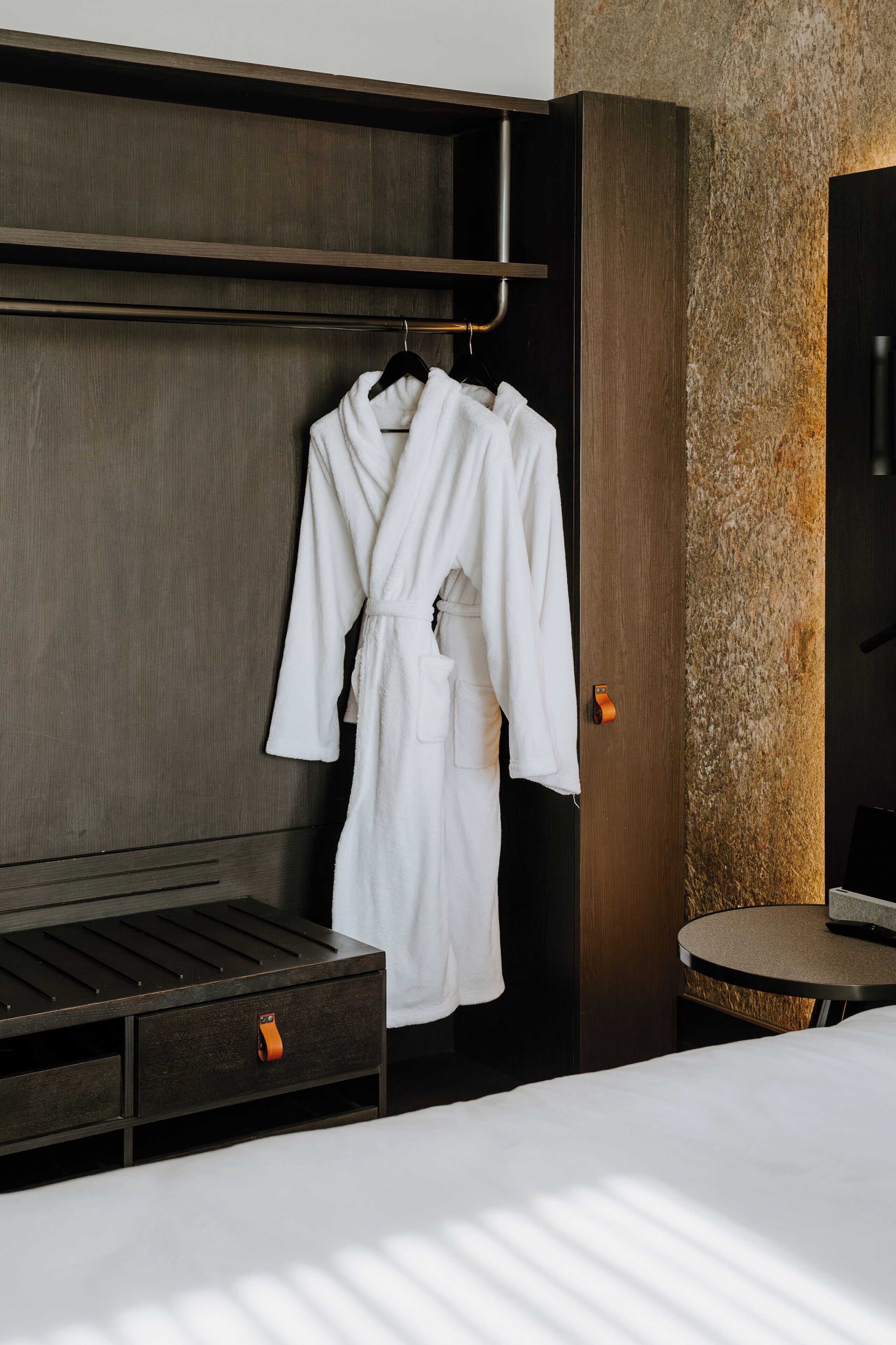Een hotelkamer met een houten garderobe, een witte badjas, een rond tafeltje en een bed met wit beddengoed.