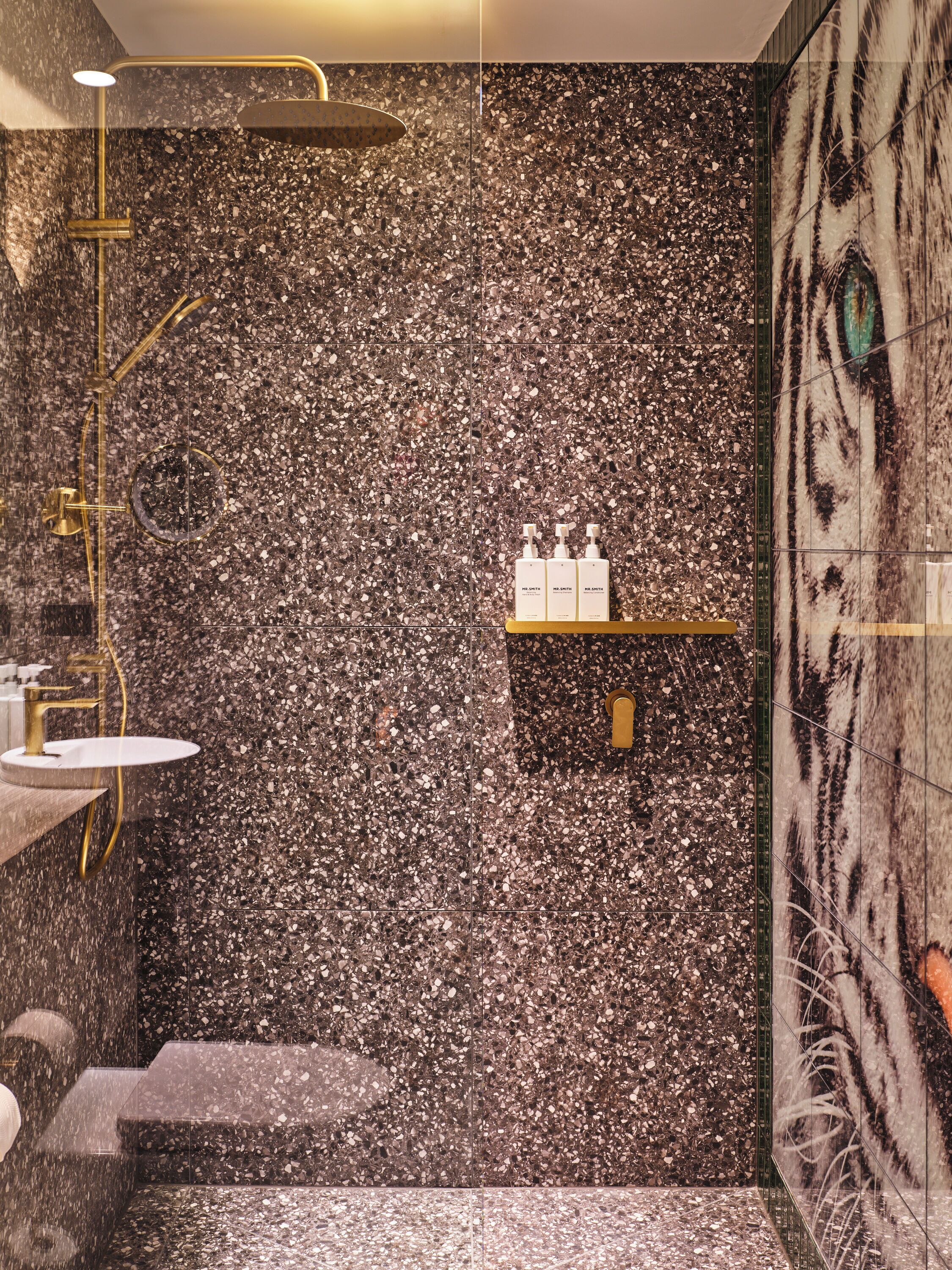 Une salle de bain moderne avec un sol en terrazzo, une pomme de douche dorée et un mur en marbre orné d’une fresque représentant un tigre.