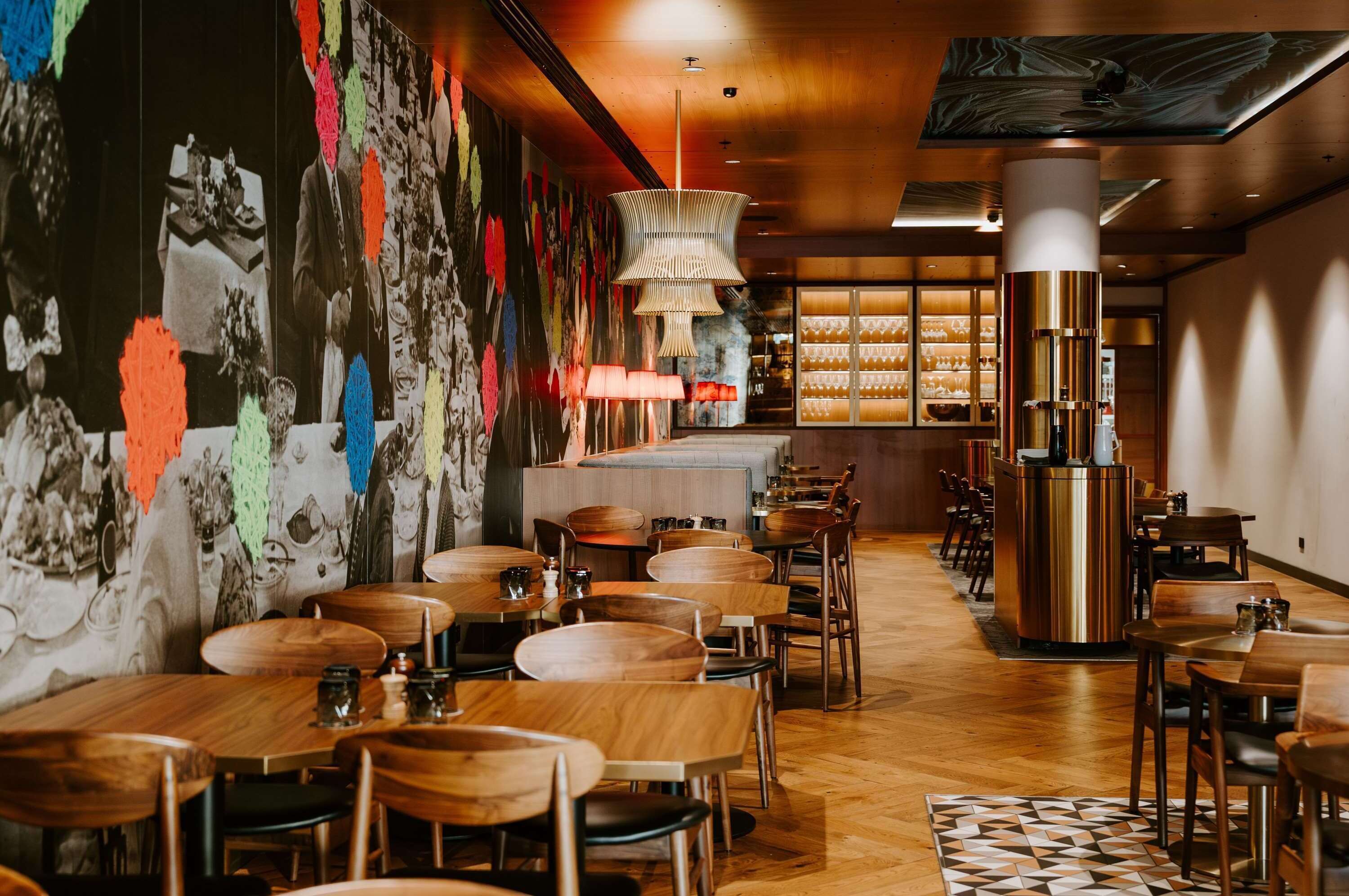 A restaurant with wooden tables and chairs, a bar area, and a wall with colorful decorations.