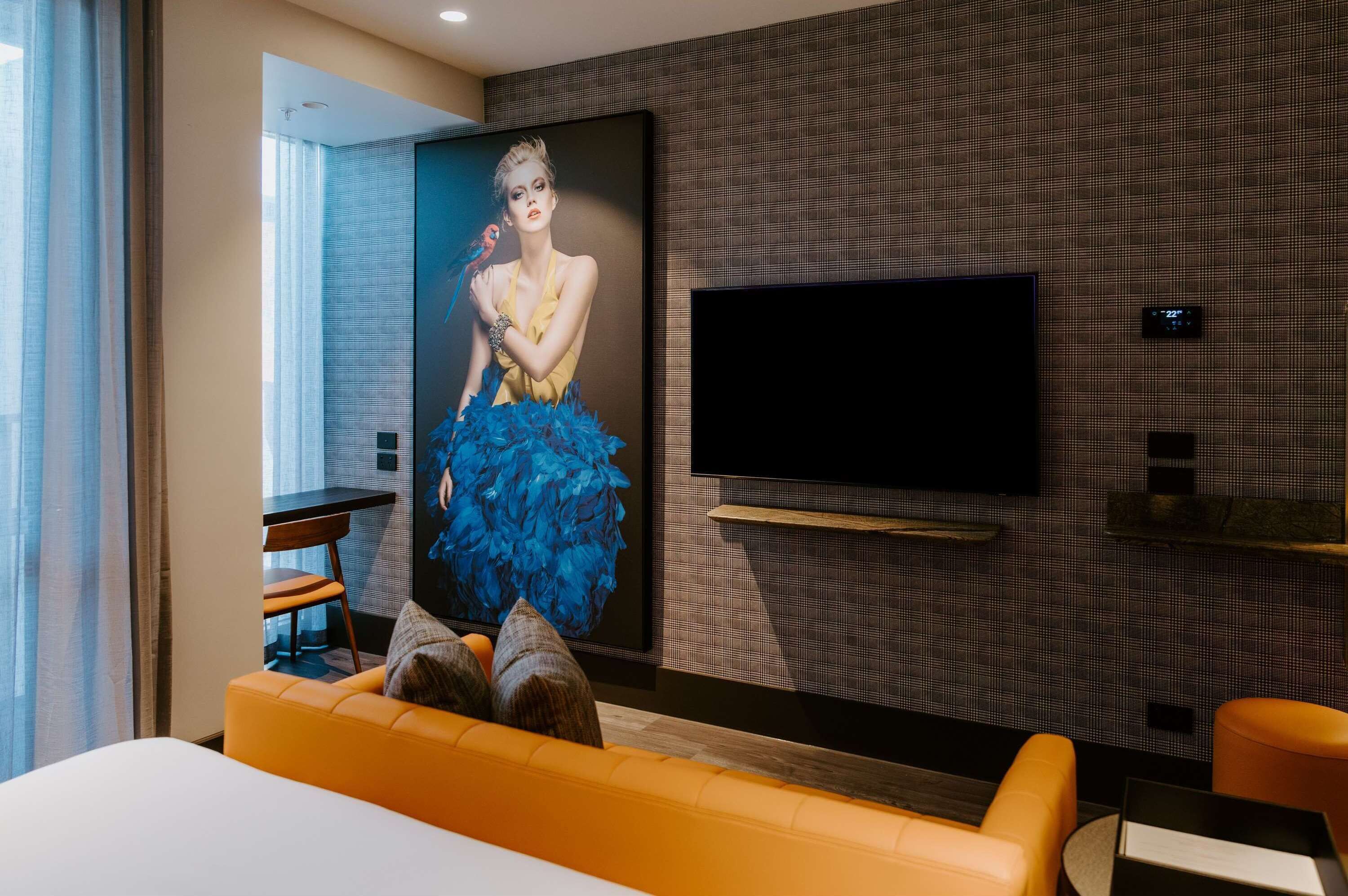 A modern hotel room with a large flat-screen TV mounted on the wall, a comfortable orange sofa, and a large poster of a woman in a blue dress.