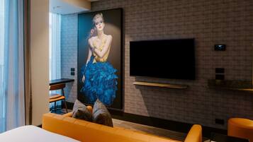Camera d'albergo moderna con una grande TV a schermo piatto montata a parete, un comodo divano arancione e un grande poster di una donna con un abito blu.