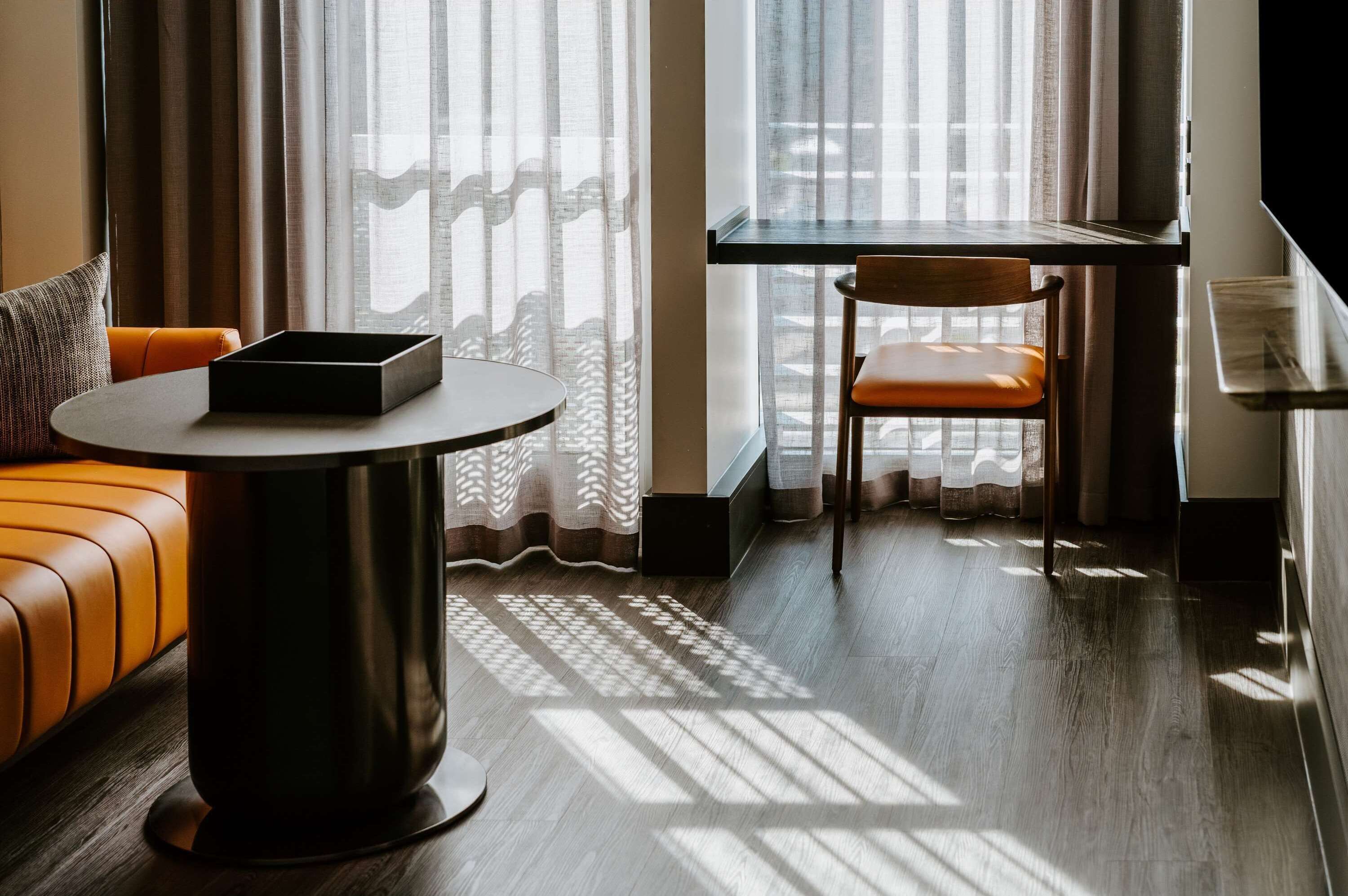 A modern hotel room with a wooden floor, a round table with a black tray, an orange leather sofa, and a wooden chair.