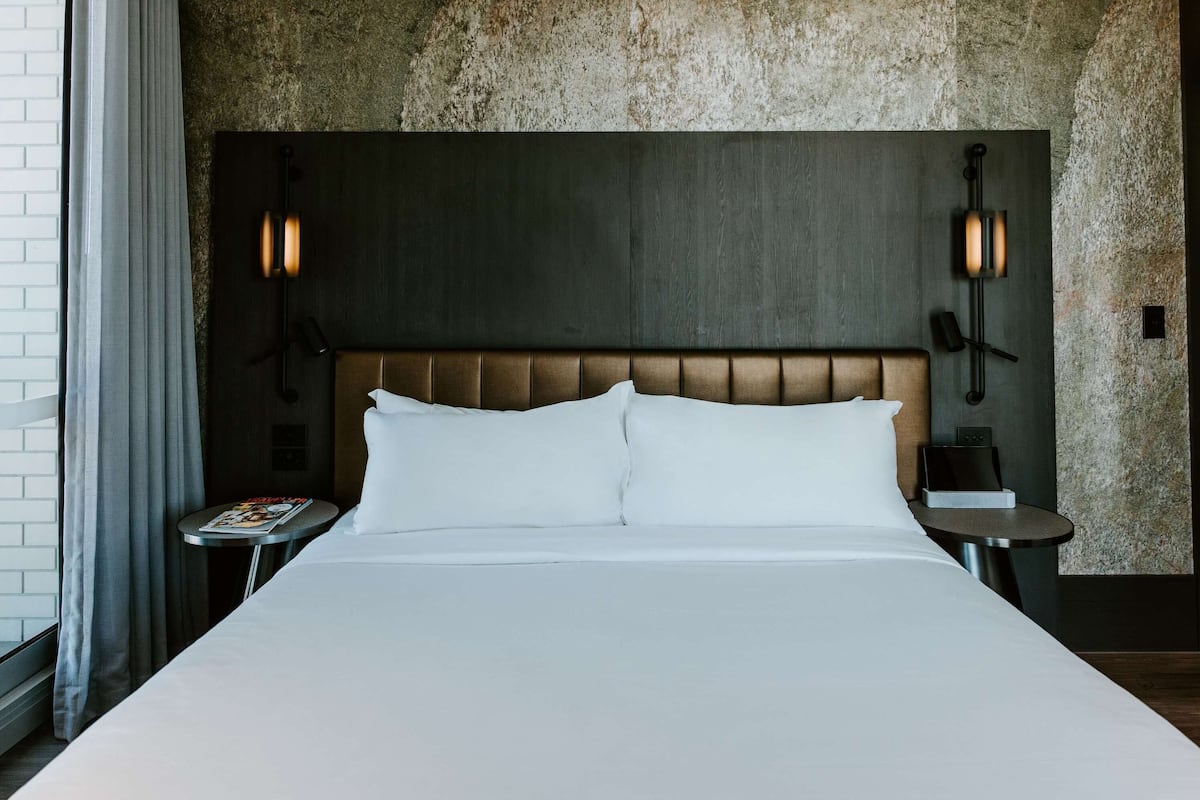 Une chambre d’hôtel avec un grand lit, une tête de lit ornée d’un panneau texturé, deux lampes de chevet, une petite table avec un magazine et une fenêtre avec des rideaux.