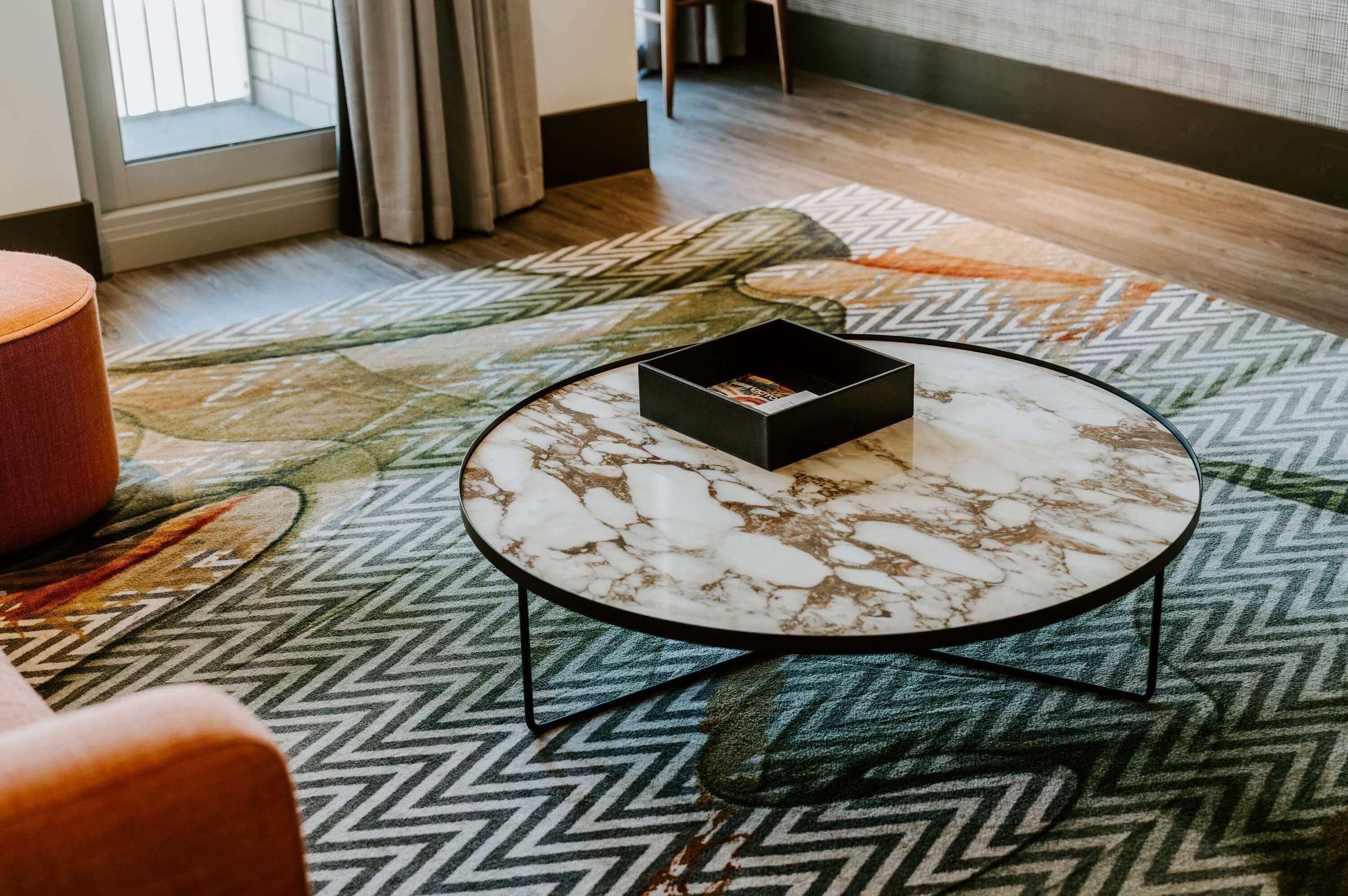 Une pièce avec un tapis à motifs, une table basse ronde en marbre sur laquelle se trouve une boîte noire, et un fauteuil.