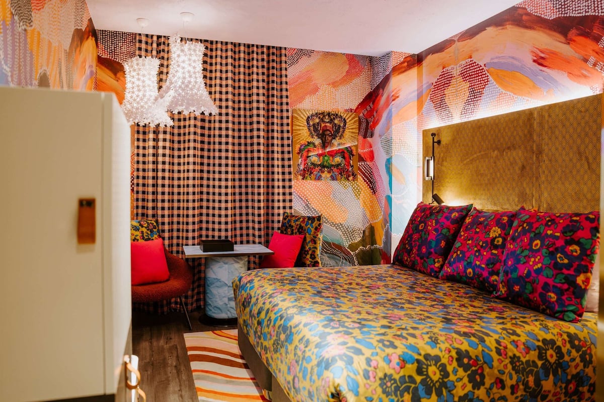 Une chambre avec un mur à carreaux, un lit recouvert d’une literie à motifs floraux colorés, une petite table avec une chaise et une décoration murale.