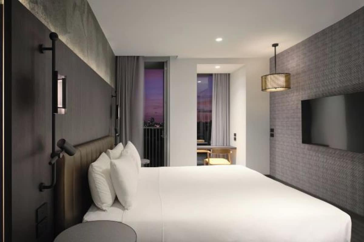 Una habitación de hotel moderna con una cama grande, una mesita de noche, un televisor montado en la pared y vistas a la ciudad a través de la ventana.