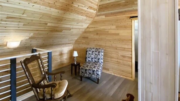 Un interno in legno con soffitto spiovente, una poltrona a dondolo, una sedia a fantasia e un tavolino con una lampada.