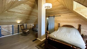 Una stanza in legno con soffitto alto, un letto, una poltrona a dondolo e un piccolo tavolo con una lampada.