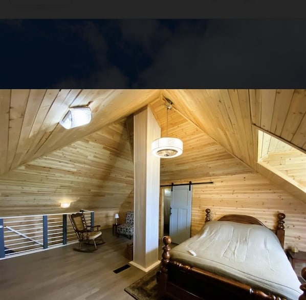 A high-ceilinged wooden room with a bed, a rocking chair, and a small table with a lamp.