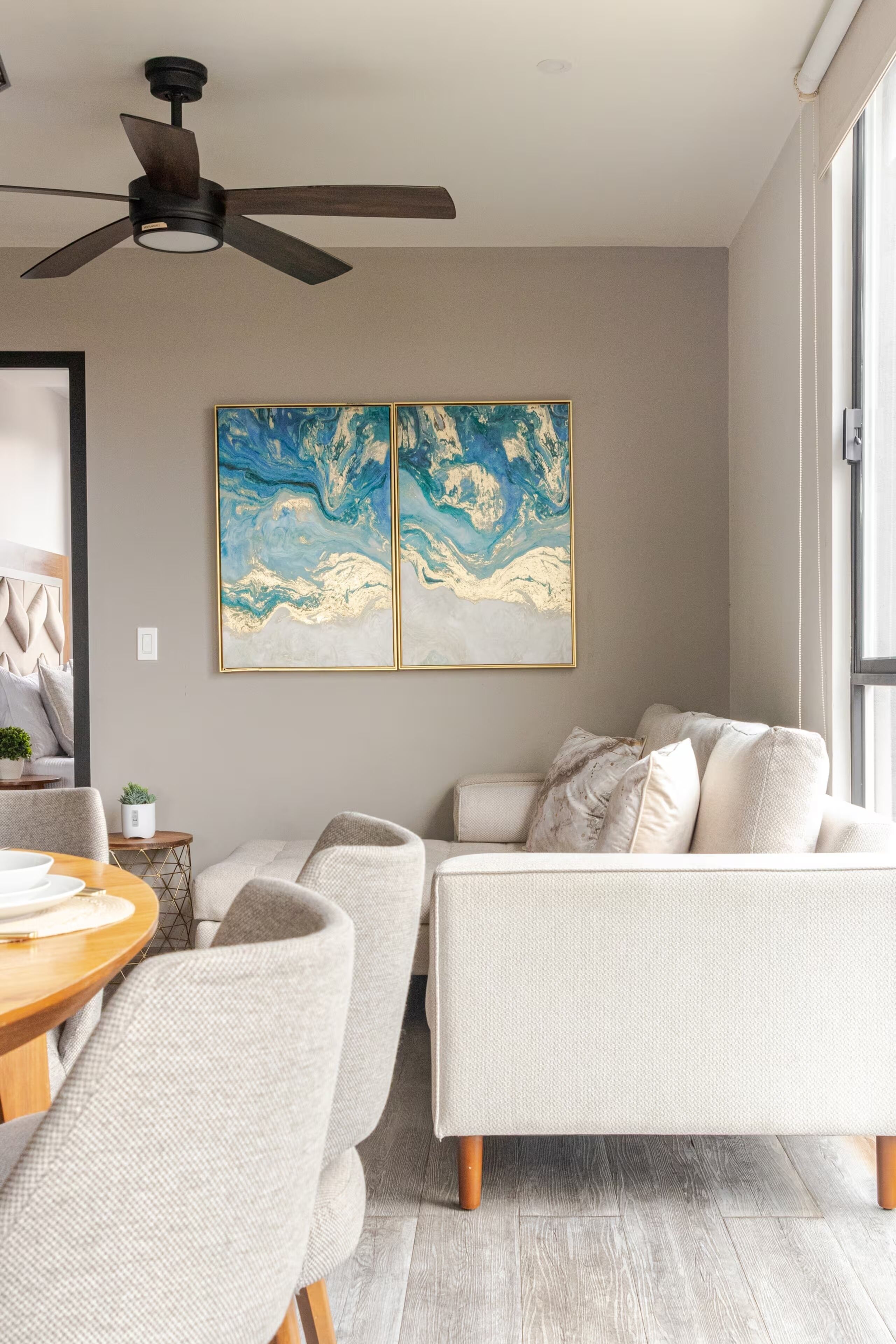 A modern living room with a ceiling fan, a dining area with a round table, and two large abstract paintings on the wall.