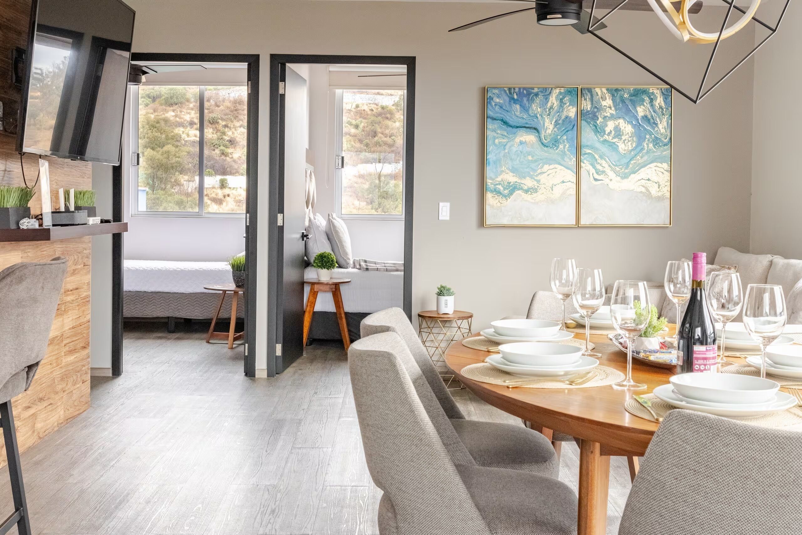 A modern dining area with a wooden table set for a meal, a large abstract painting on the wall, and a view of the outdoors through a glass door.