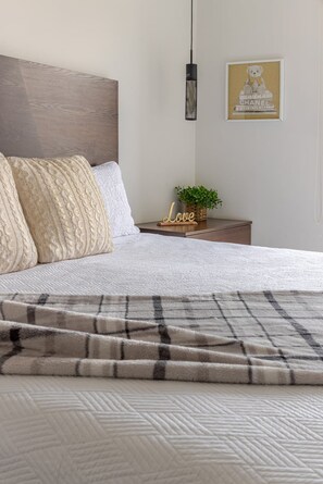 A neatly made bed with a patterned blanket, a bedside table with a plant and a 'Love' sign, and a framed picture on the wall.