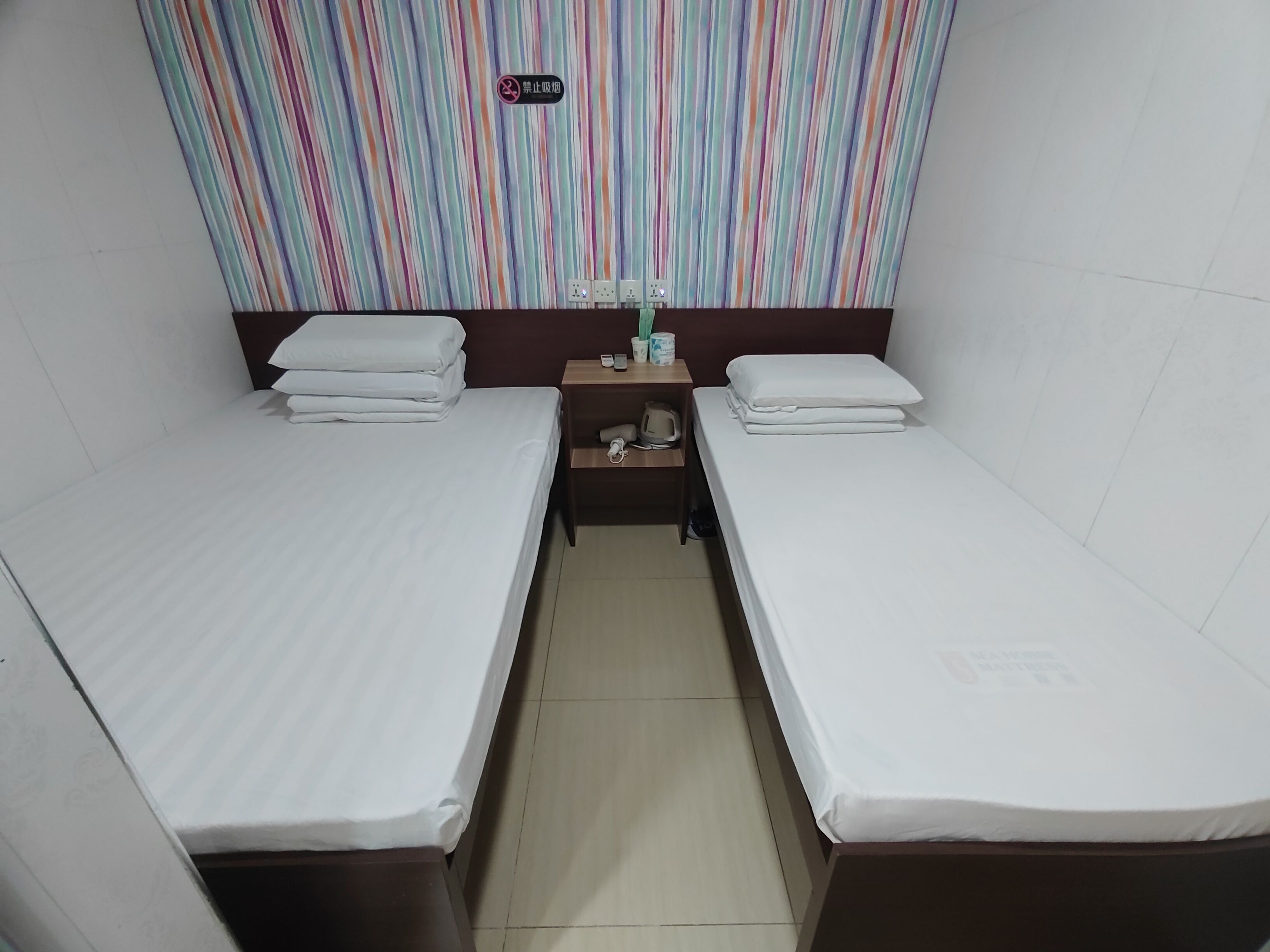 A small, clean room with two single beds, a nightstand with a phone and a cup, and a striped curtain.