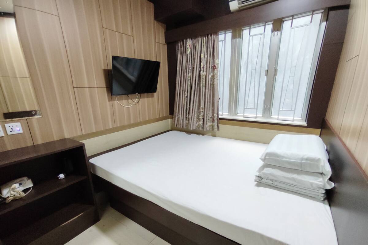 A hotel room with a bed, a TV mounted on the wall, a window with curtains, and a small shelf with a phone.
