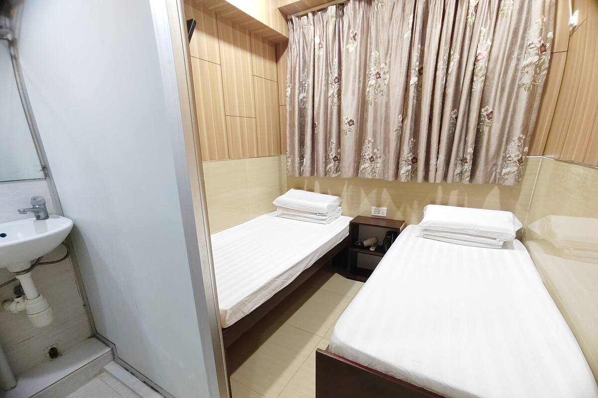 A small, clean room with two single beds, a bedside table, a nightstand, a small shelf with a lamp, and a bathroom with a sink and mirror.
