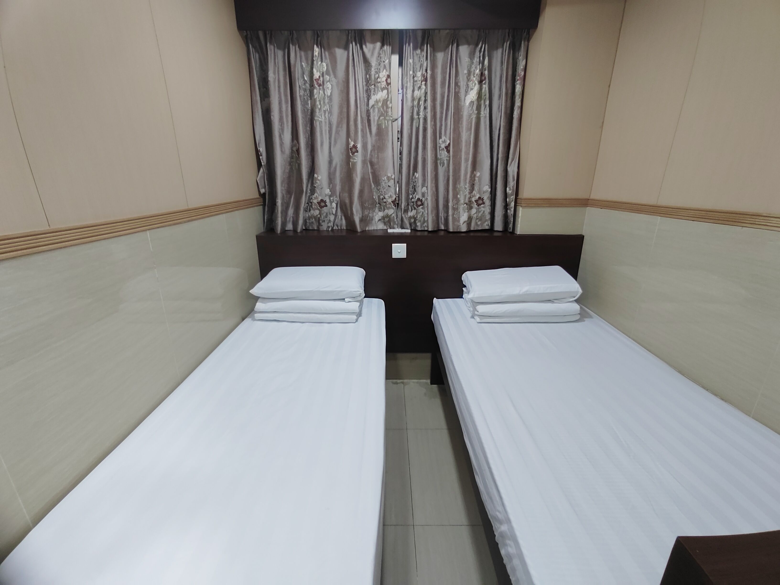 A hotel room with two single beds, a wooden headboard, a curtain with a floral pattern, and a light-colored tiled floor.
