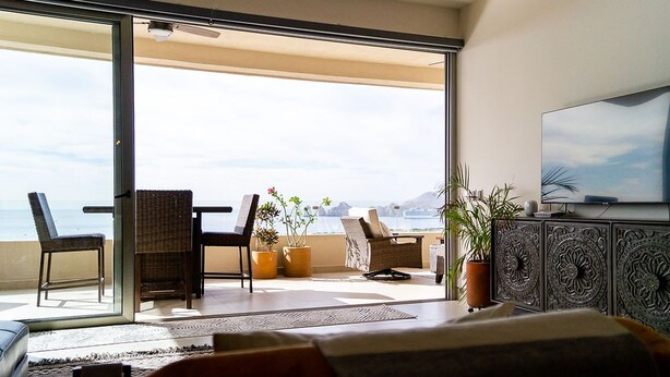 A balcony with a view of the sea, furnished with chairs and a table, and a TV on a cabinet. - Fabulous Cabo condo with amazing view of lands end! (Cabo San Lucas)