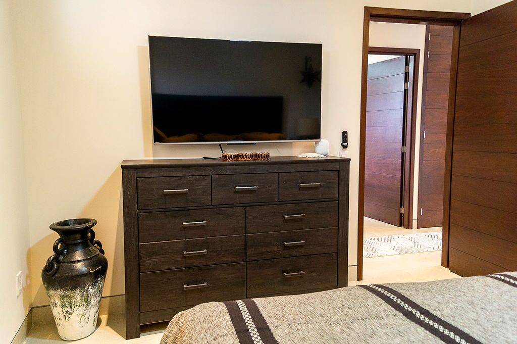 A bedroom with a large flat-screen TV mounted on a dark wooden dresser, a decorative vase, and a bed with a patterned blanket.