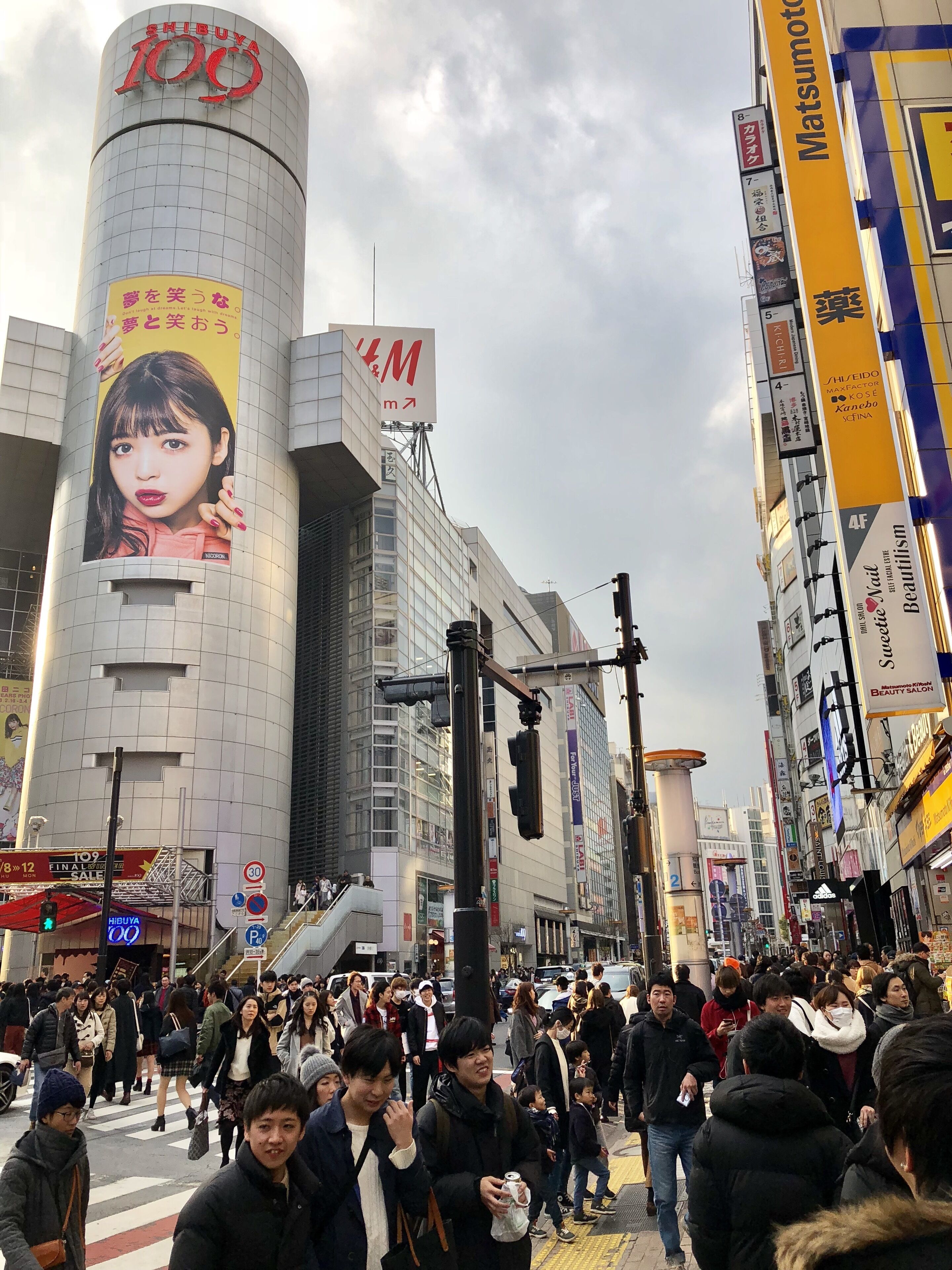 Eine belebte Stadtstraße mit einem markanten Gebäude, das eine große Werbetafel und Schilder für Shibuya 109 zeigt.