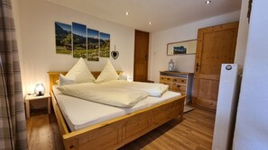 A wooden bed with white bedding, a nightstand with a lamp, a dresser with drawers, a mirror, and a door with a wooden panel.