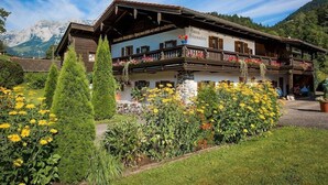 Een traditioneel houten Alpengebouw met balkon, omgeven door verzorgde tuinen en bergen op de achtergrond.