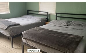 Two single beds with wooden frames and grey bedding, situated in a room with a green wall and a window with blinds.