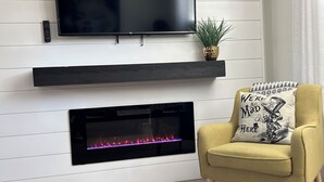 Ein modernes Wohnzimmer mit Flachbildfernseher über einem Kamin, einem gelben Sessel mit Zierkissen und einem kleinen Topfpflanzen auf dem Kaminvorsatz.