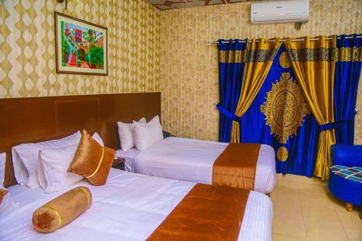Habitación de hotel con dos camas, un sillón azul y una cortina decorativa con un emblema dorado.