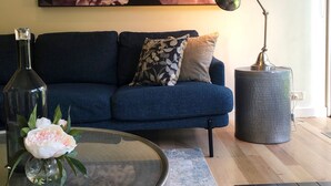 A living room with a blue sofa, a glass coffee table, a lamp, and a large floral painting on the wall.