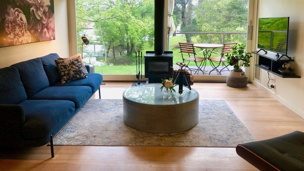 A modern living room with a blue sofa, a glass coffee table, a fireplace, and a view of a garden through large windows.