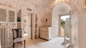 Quarto com paredes de pedra, um radiador, uma cadeira de madeira, um armário branco e uma porta que dá para o exterior.