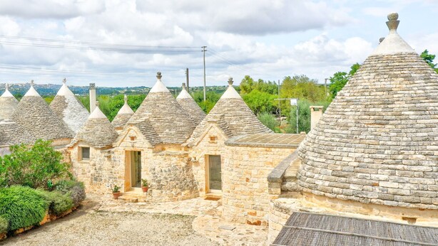 Uma fileira de edifícios com telhados de pedra, pequenas janelas e portas, cercada por vegetação.