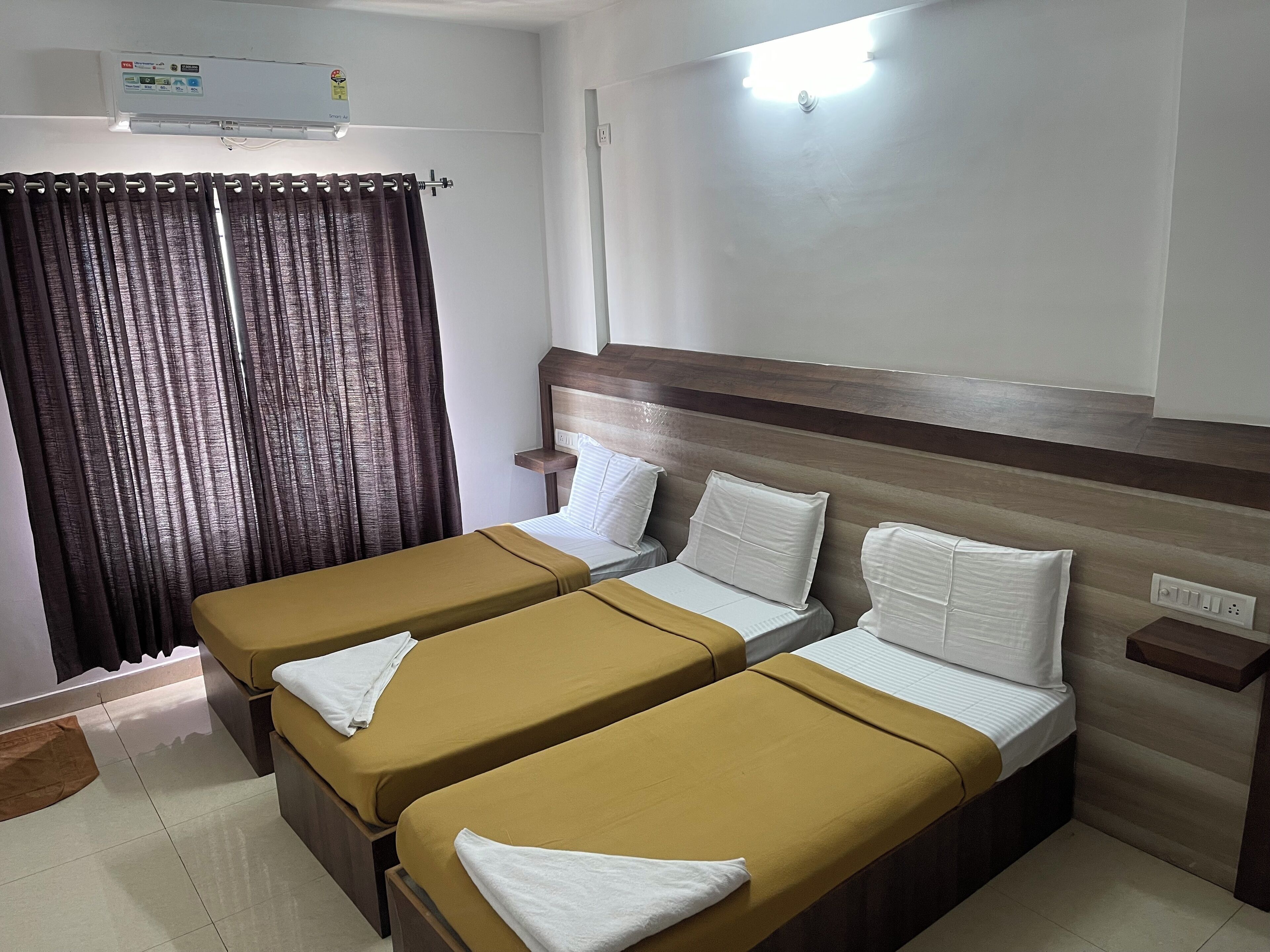 A hotel room with two single beds, a wooden headboard, a small bedside table, a wall-mounted air conditioner, and a curtain.