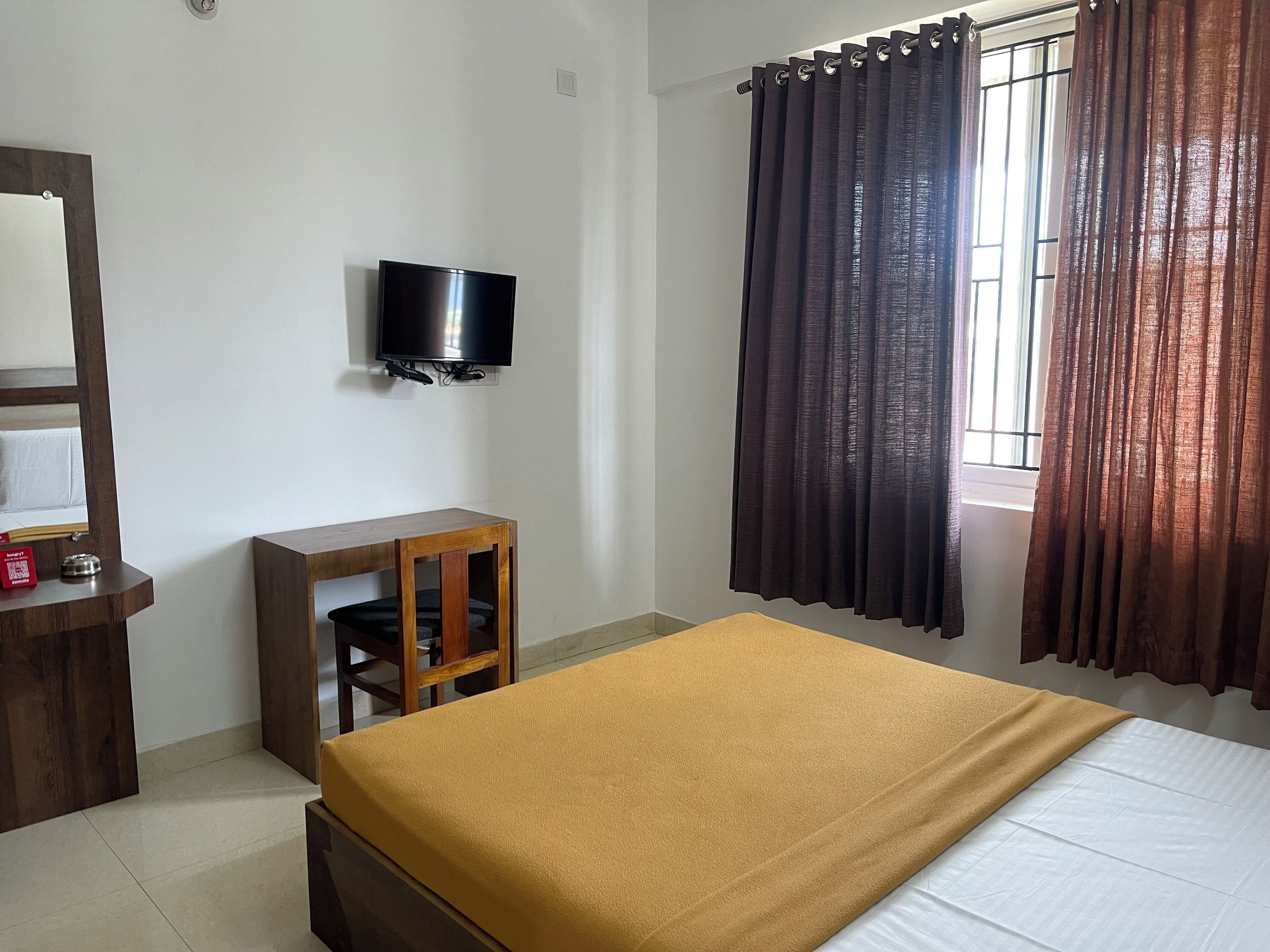 A hotel room with a bed, a desk with a chair, a television mounted on the wall, and a window with curtains.