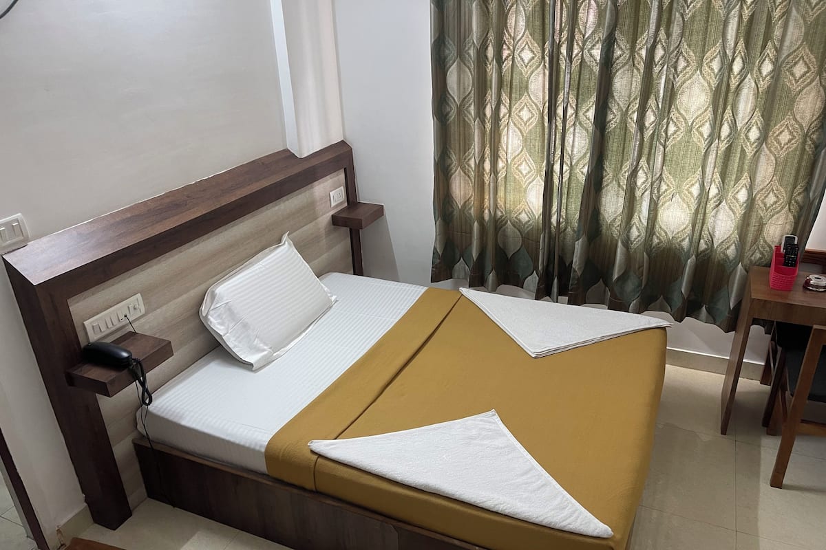 A neatly made bed with a brown bedspread and a white pillow, a bedside table with a phone and charger, and a curtain with a leaf pattern.