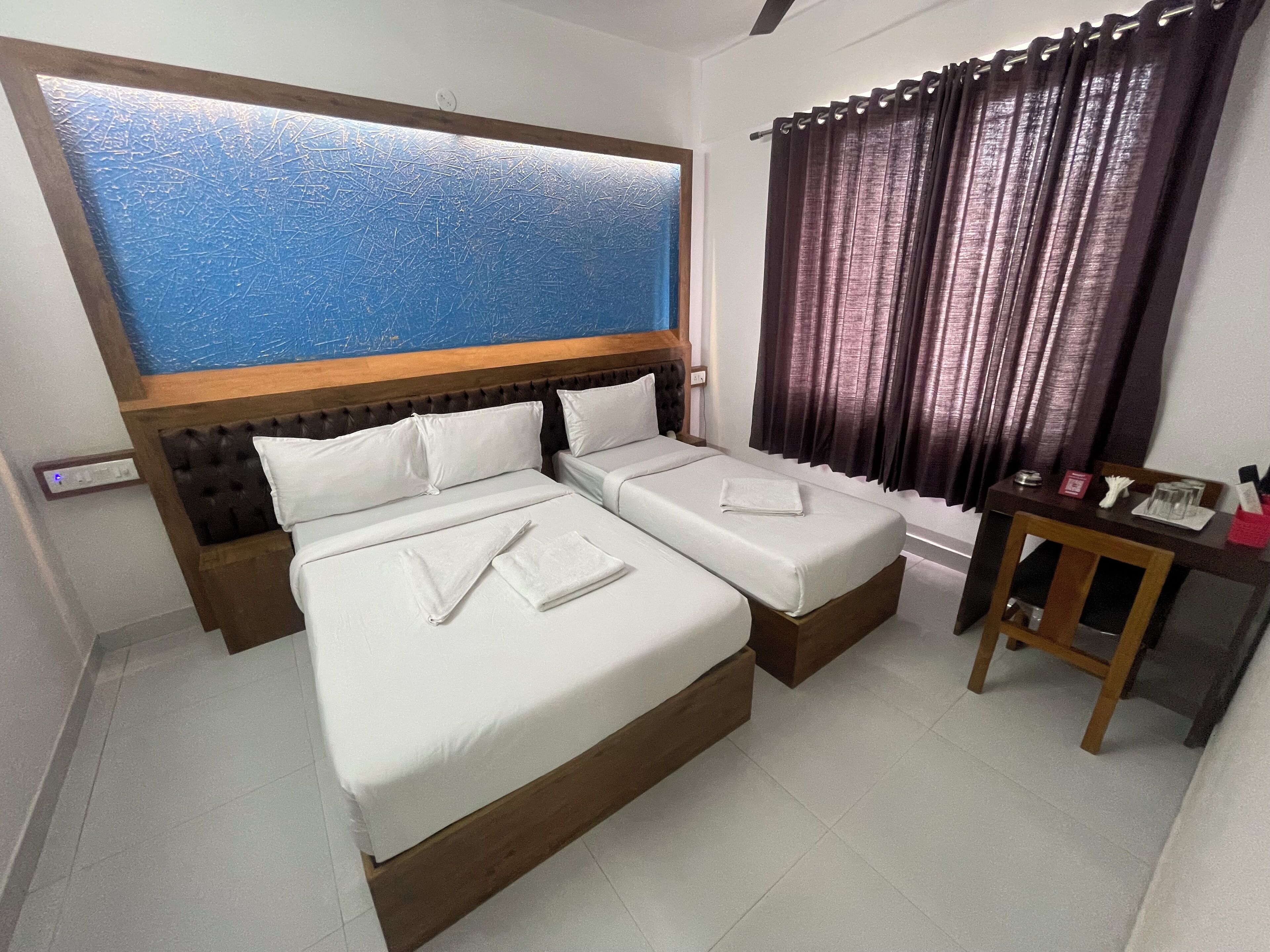 A hotel room with two beds, a wooden headboard, a small table with a chair, and a window with curtains.