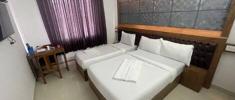 A hotel room with two beds, a wooden headboard, a small table with a chair, a TV, and a window with curtains.