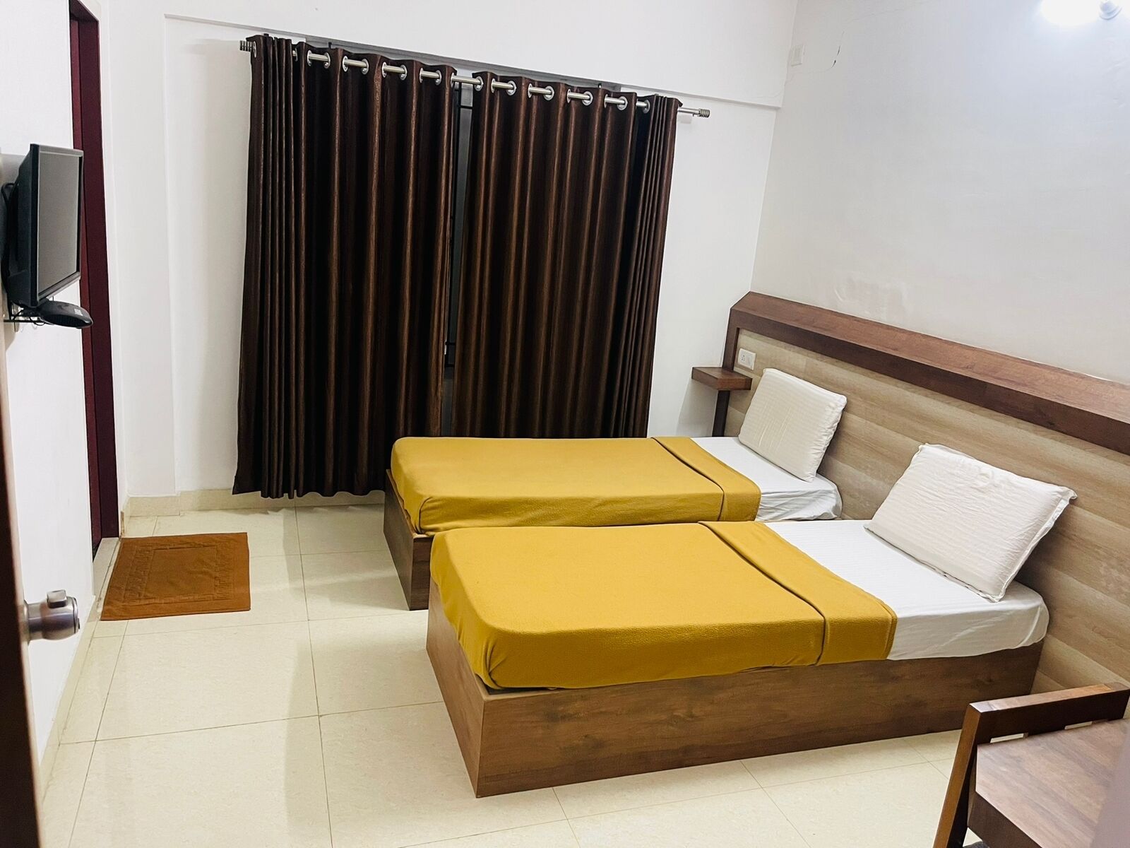 A hotel room with two single beds, a wooden headboard, a flat-screen TV mounted on the wall, and a curtain covering the window.