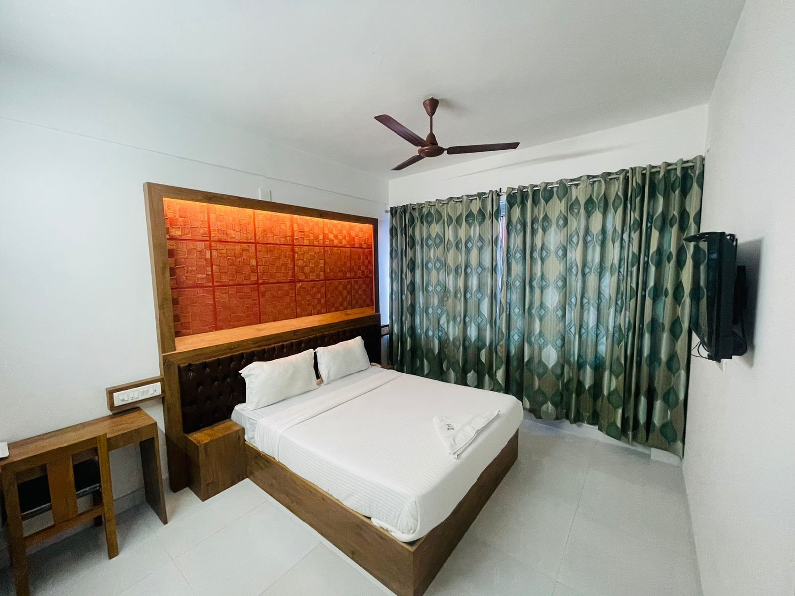 A bedroom with a bed, a wooden headboard, a ceiling fan, a TV mounted on the wall, and a window with patterned curtains.