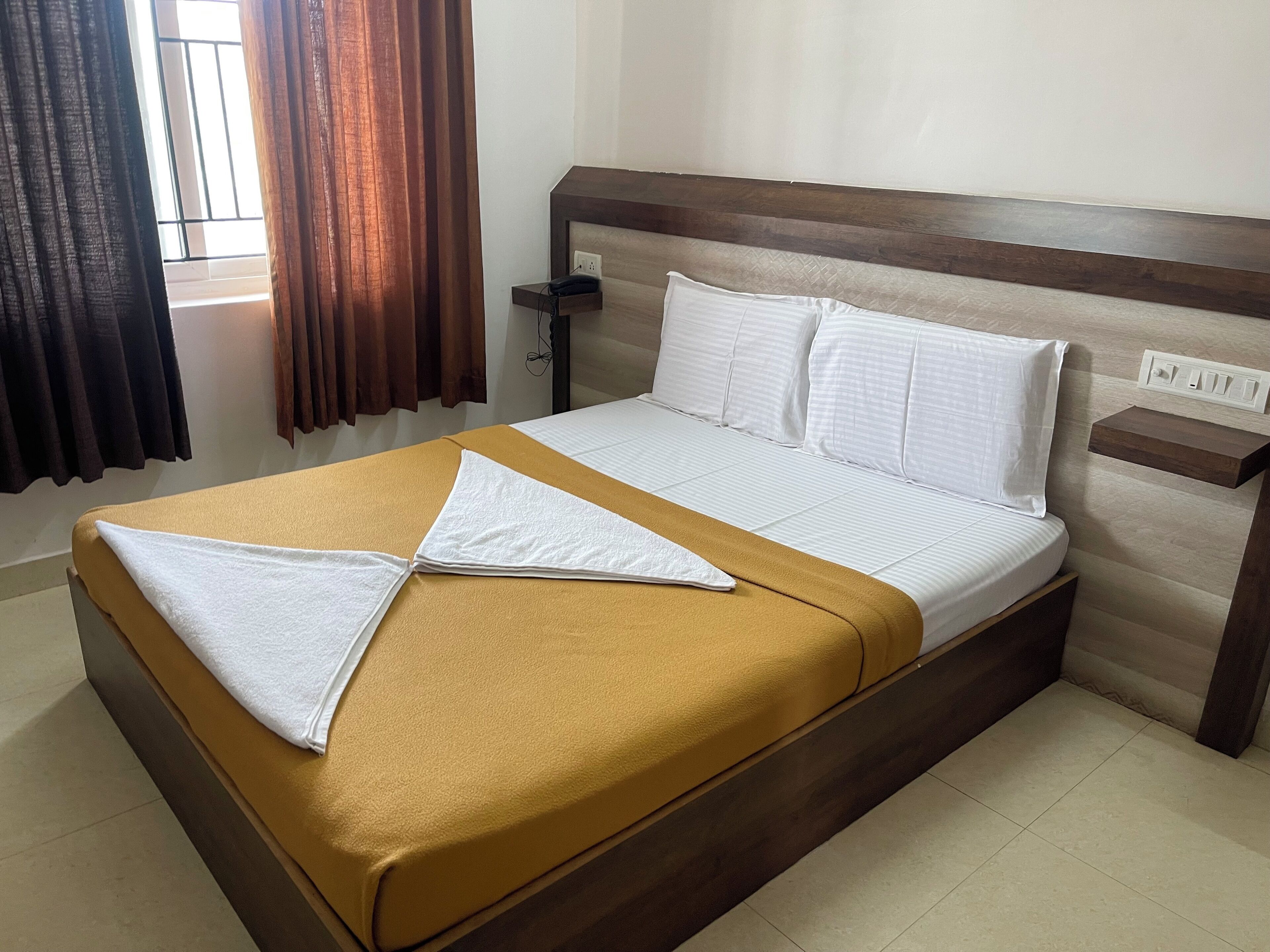 A neatly made bed with a brown bedspread and white pillows, a wooden headboard, a bedside table with a lamp, and a window with curtains.