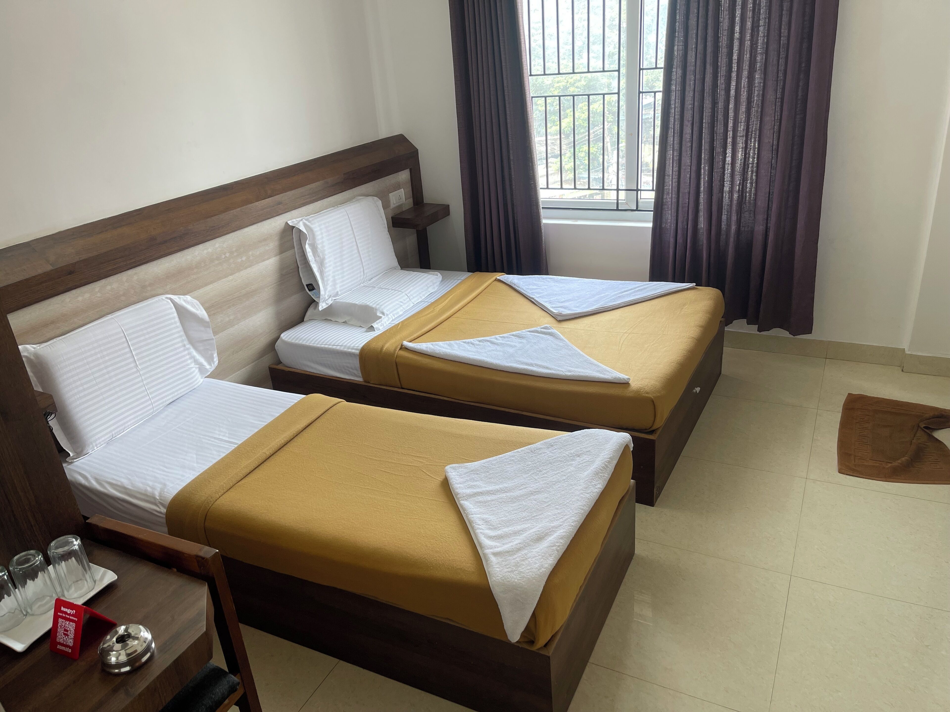 Quarto de hotel com duas camas, uma mesinha com um tabuleiro de copos e uma janela com cortinas.